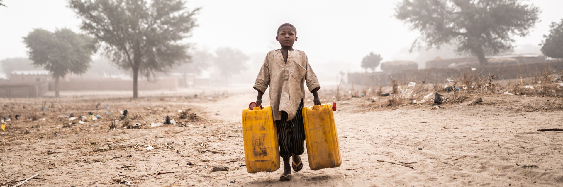 Kind trägt in einer ausgedörrten Landschaft zwei Kanister gefüllt mit Wasser.