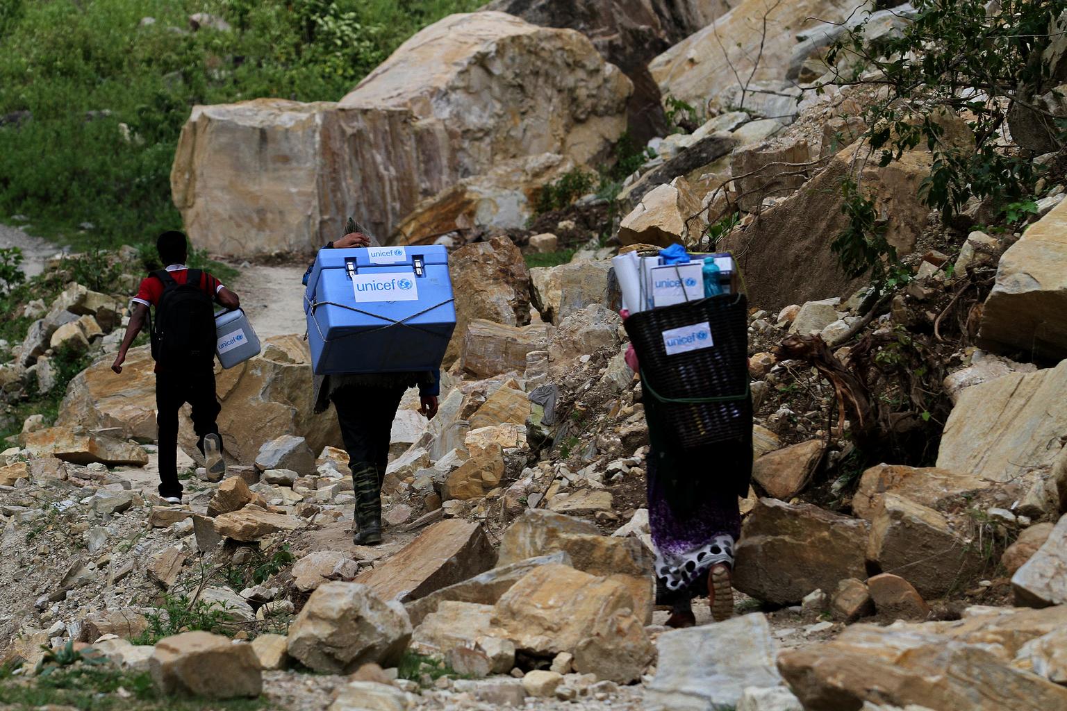 Drei Helfer haben Kühlboxen am Rücken und klettern zu einem Dorf.