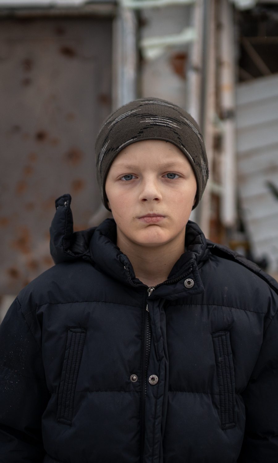 Junge in Winterkleidung steht vor einem zerstörten Haus in der Ukraine.